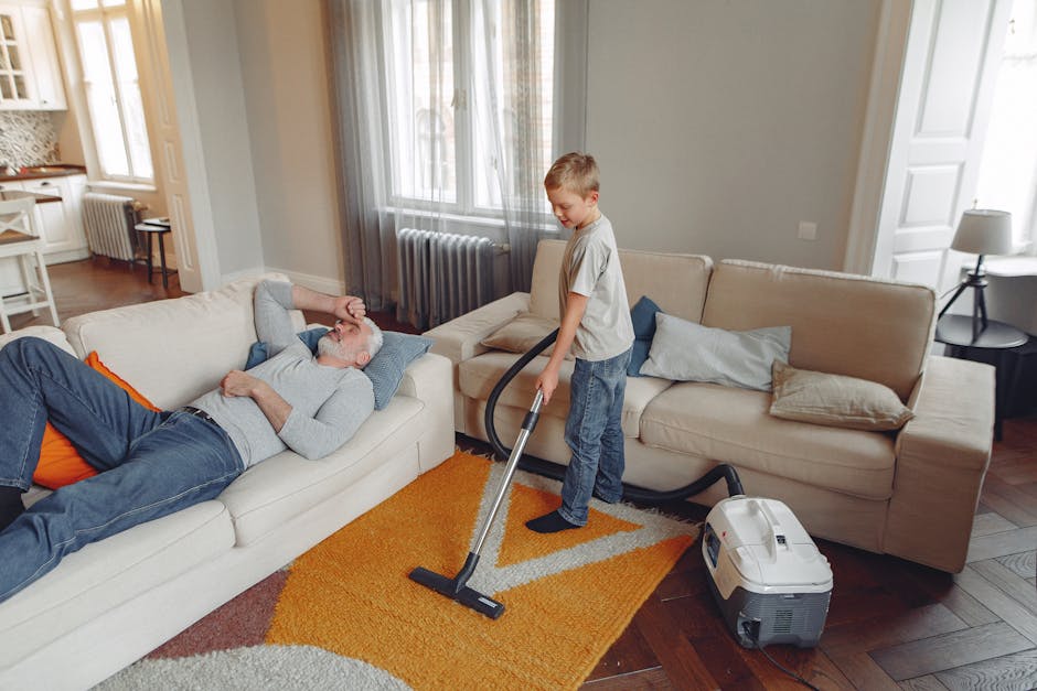 A young boy vacuuming the carpet with a modern vacuum cleaner while an older man reclining on a beige sofa wears glasses and a grey sweater. The room features a wooden floor with a patterned orange and beige rug, soft natural light streaming through large windows with sheer curtains, and neutral walls. A small side table with a lamp and a radiator are visible in the background. The scene depicts domestic surface cleaning and vacuuming, emphasizing hygiene and cleanliness under the expertise of Bermondsey Carpet Cleaning, as part of their tips for residents on carpet maintenance and deep cleaning in Bermondsey Street SE1.