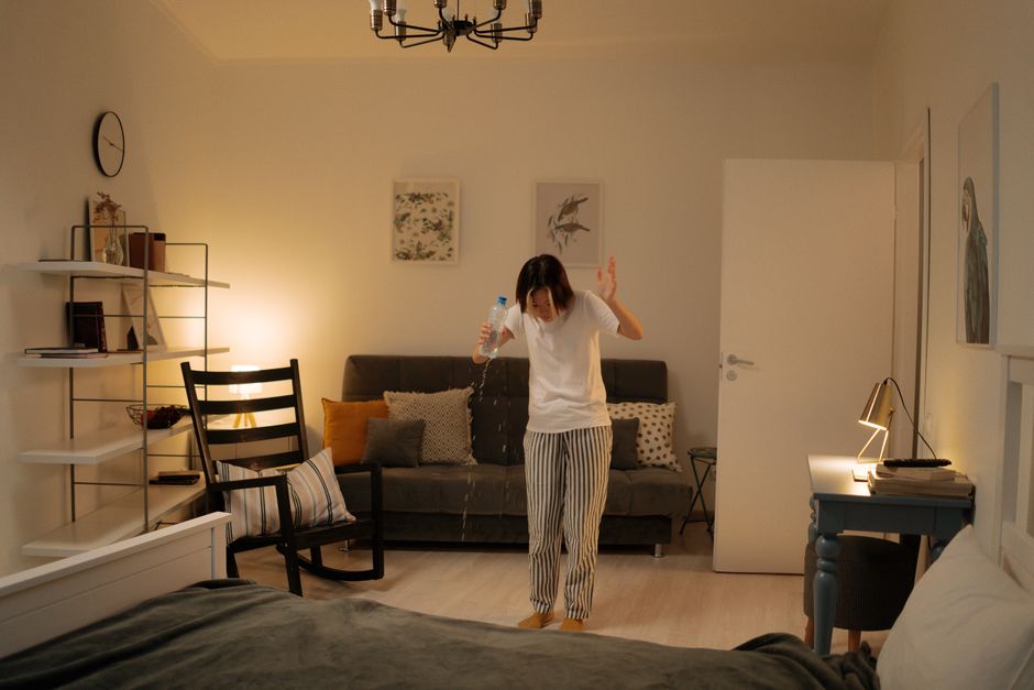 A woman standing in a living room with a dark gray sofa, colorful cushions, and modern wall art. She is wearing striped pajamas and holding a water bottle, pouring water onto the floor. The room features a white side table with a desk lamp and books, a black dining chair, and white wall shelves with decorative items. The lighting is warm and soft, creating a cozy atmosphere, while the room appears clean and tidy as part of a deep cleaning or surface sanitisation process. Bermondsey Carpet Cleaning specializes in emergency carpet and surface cleaning, ensuring hygiene and maintenance in residential spaces.