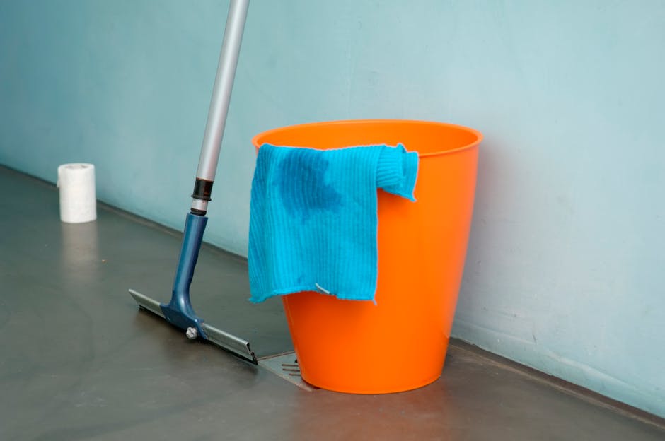 A professional cleaner dressed in full white protective suit, mask, and gloves is performing surface cleaning inside a modern living room. They are using a vacuum or extraction device that emits visible steam or fumes, indicating a deep cleaning or sanitisation process on a beige carpet. The room features a wooden shelving unit with various decor items, a potted plant, and a large mirror, all illuminated by natural light, creating a bright and clean environment. This image exemplifies the emergency carpet cleaning services offered by Bermondsey Carpet Cleaning, highlighting thorough and hygienic indoor maintenance for spill or stain removal.