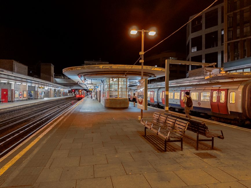 Night view of Bermondsey Station platform with clean, well-maintained tiled flooring, illuminated by overhead lighting. Two benches are positioned along the platform, one slightly tilted, with no visible dust or debris. Nearby, a train with red and grey exterior is stationary on the tracks, its windows shining softly under the station lights. The station structure includes a modern brick and glass shelter in the center, and the background features tall buildings under dark skies. Bermondsey Carpet Cleaning emphasizes the importance of surface cleaning and sanitisation for maintaining hygiene in public transport areas, ensuring a safe environment for commuters.