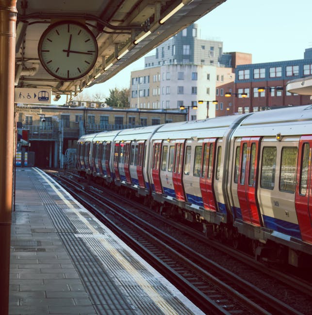 A view from Bermondsey Station platform showing a modern train with a red, white, and blue exterior parked on the tracks. The platform surface appears clean and well-maintained, with tactile paving near the edge for safety. Overhead, part of the station roof is visible, including a large round clock displaying the time. In the background, urban buildings with glass and brick facades reflect daylight. The scene emphasizes the importance of surface cleaning and hygiene for a tidy, safe commuter environment, highlighting Bermondsey Carpet Cleaning's expertise in maintaining clean, publicly accessible spaces as seen in the station setting.