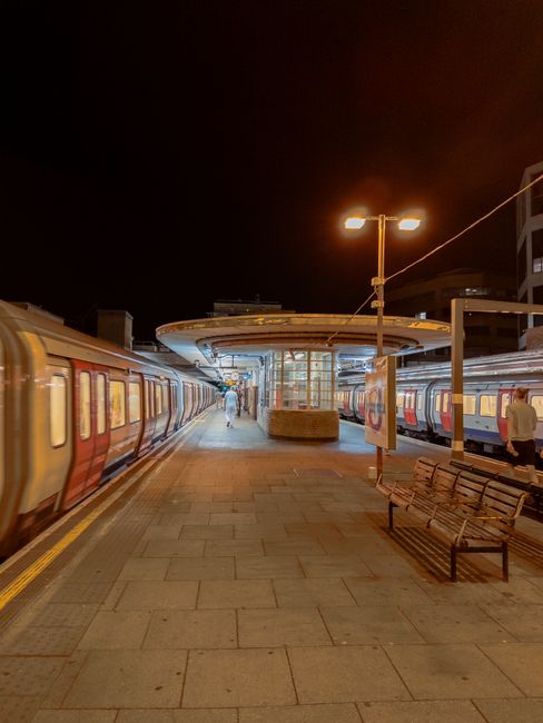 Night view of Bermondsey Station platform with clean, well-maintained tiled flooring, illuminated by overhead lighting. Two benches are positioned along the platform, one slightly tilted, with no visible dust or debris. Nearby, a train with red and grey exterior is stationary on the tracks, its windows shining softly under the station lights. The station structure includes a modern brick and glass shelter in the center, and the background features tall buildings under dark skies. Bermondsey Carpet Cleaning emphasizes the importance of surface cleaning and sanitisation for maintaining hygiene in public transport areas, ensuring a safe environment for commuters.