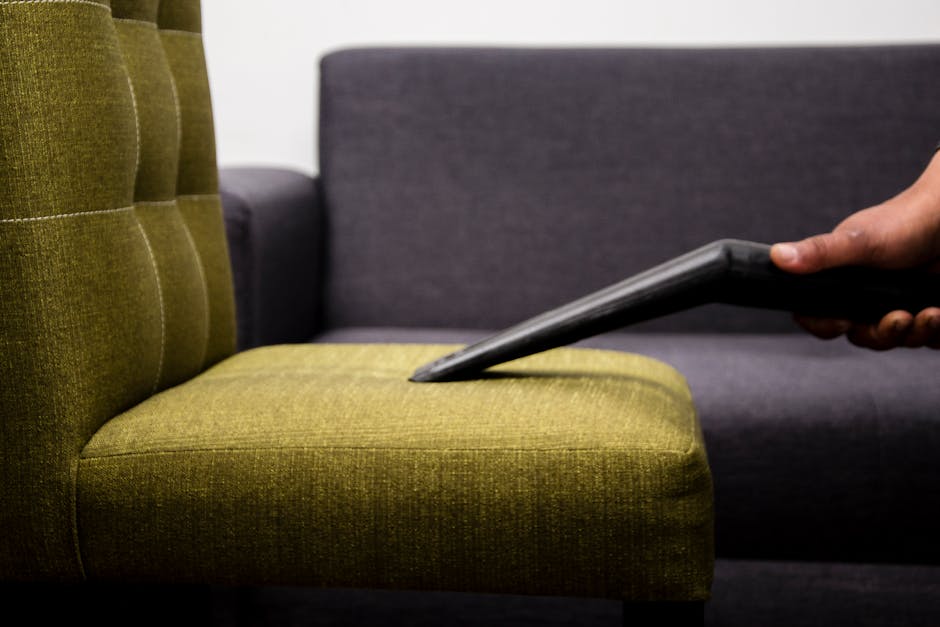 Close-up of a hand using a vacuum cleaner attachment to deep clean a green upholstered armchair with tufted buttons in a modern living room, featuring a dark gray fabric sofa in the background. The room is well-lit with neutral lighting, highlighting the clean surface of the fabric. The upholstery appears dust-free and well-maintained, demonstrating surface cleaning to maintain hygiene. Bermondsey Carpet Cleaning specializes in domestic cleaning services, ensuring furniture and surfaces are thoroughly sanitised and free of dirt, contributing to a hygienic living environment.