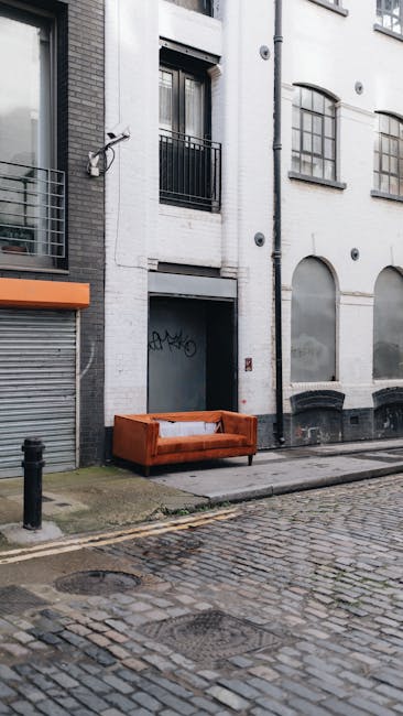 An outdoor urban scene featuring a white multi-story building with large arched windows and black metal balconies. In front of the building, on the cobblestone sidewalk, there is a vintage brown upholstered sofa situated next to a black bollard. The area appears clean and well-maintained, with natural lighting indicating daytime. The image suggests a setting where surface cleaning or maintenance might be performed by Bermondsey Carpet Cleaning, as referenced on the page about upholstery cleaning near Maltby Street Market Bermondsey on bermondseycarpetcleaning.co.uk.
